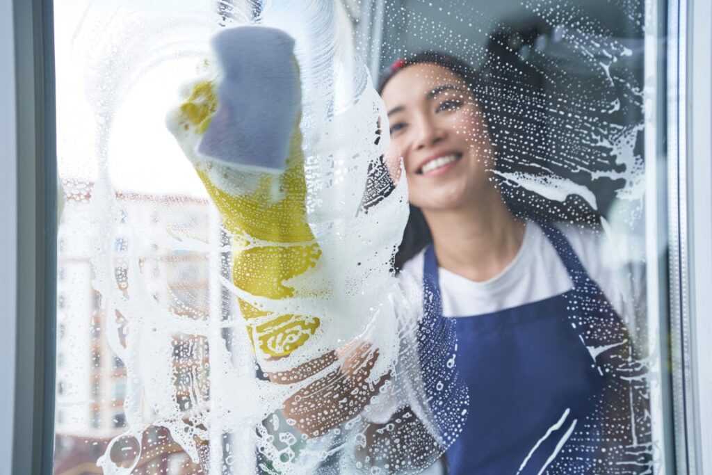 Best cleaning service. Cheerful young woman smiling while cleaning the window, glass surface using sponge