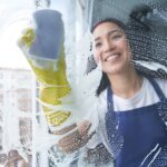 Best cleaning service. Cheerful young woman smiling while cleaning the window, glass surface using sponge
